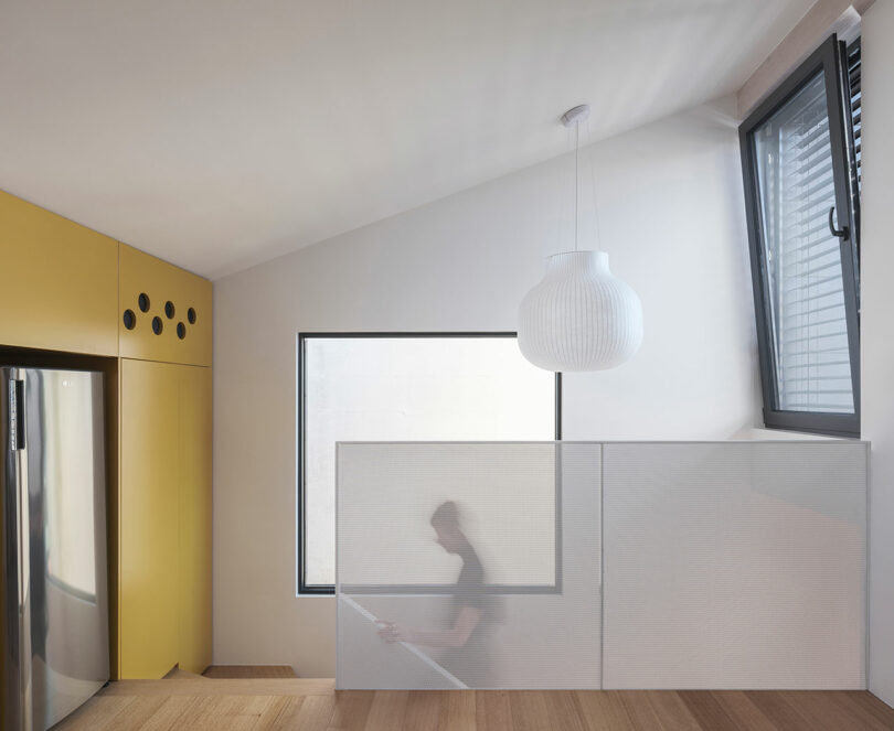 Modern interior with yellow cabinets, large stainless steel refrigerator and a blurred person in motion behind a semi-transparent partition, near a window with a geometric lamp suspended from the ceiling.