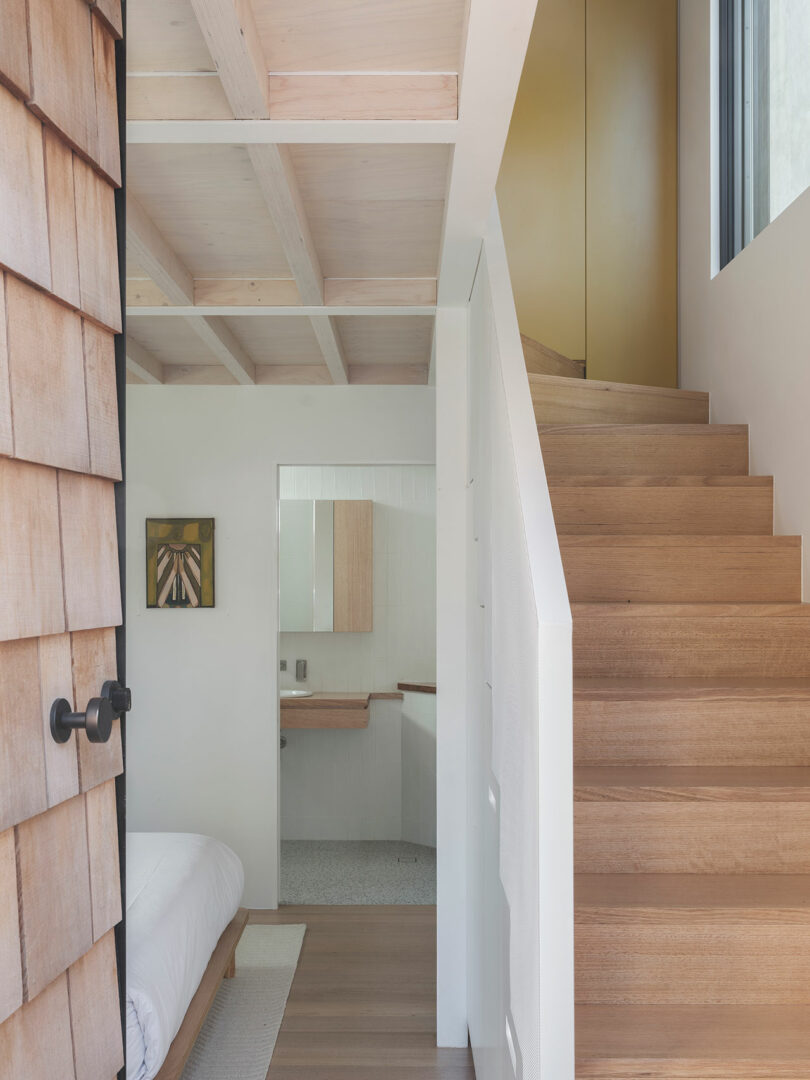 Interior showing a wooden staircase leading to the upper level, a bedroom with a white bed and a partial view of a bathroom with a mirror and artwork on the wall.