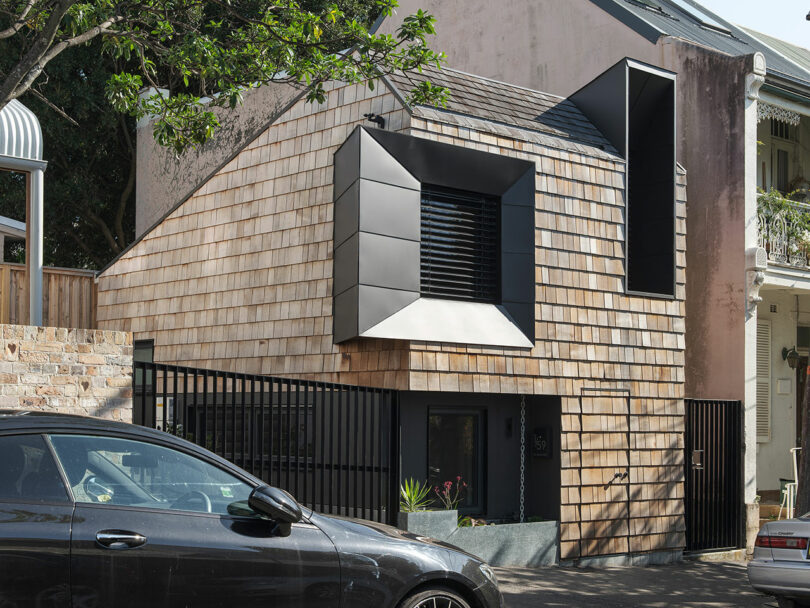 Modern house with tile siding and large geometric window, with a black fence in front and a parked car.