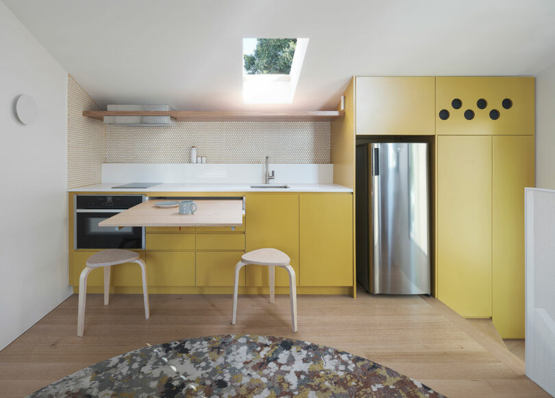 Modern kitchen with yellow cabinets, built-in oven, stainless steel refrigerator and two wooden benches. A skylight provides natural light. The walls feature a white tile backsplash.