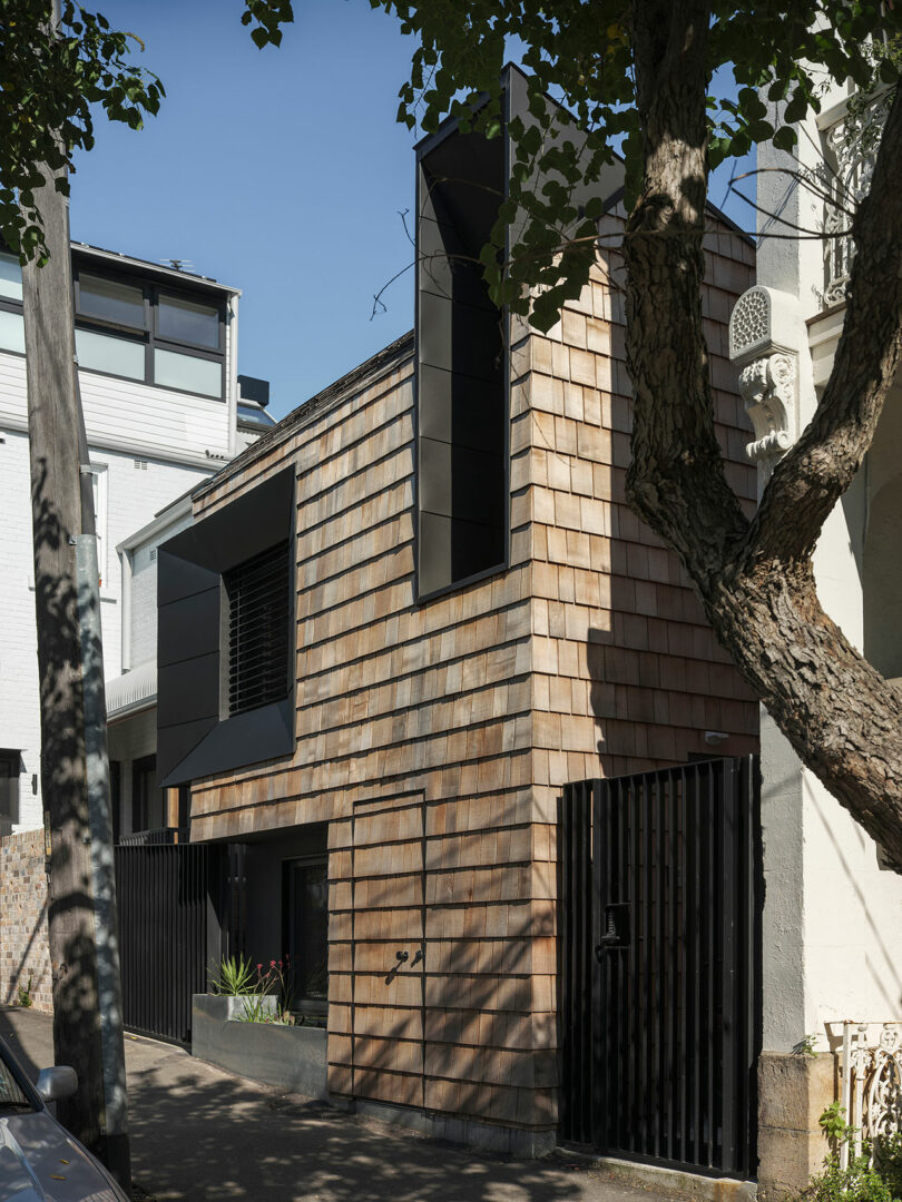 Modern three-story house with a wooden shingle facade, large black-framed windows and a tree in front. The house is flanked by a gate and neighboring buildings.